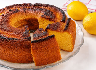 Bolo de Limão Simples e Muito Fácil de Fazer Bolo de limão simples, alto, fofinho e dourado, com uma fatia cortada que revela um interior macio e húmido, servido numa boleira de vidro com limões frescos ao fundo.